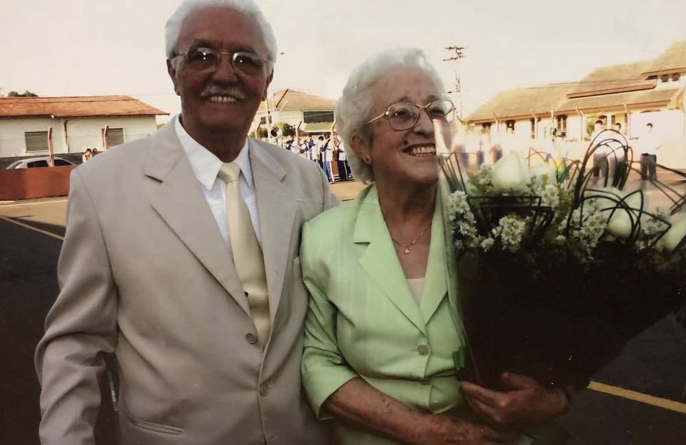 Seu Barros e Dona Rony Fecci Piekarski, sorridentes, em uma solenidade da Guarda Mirim; ela segura um buquê de flores brancas